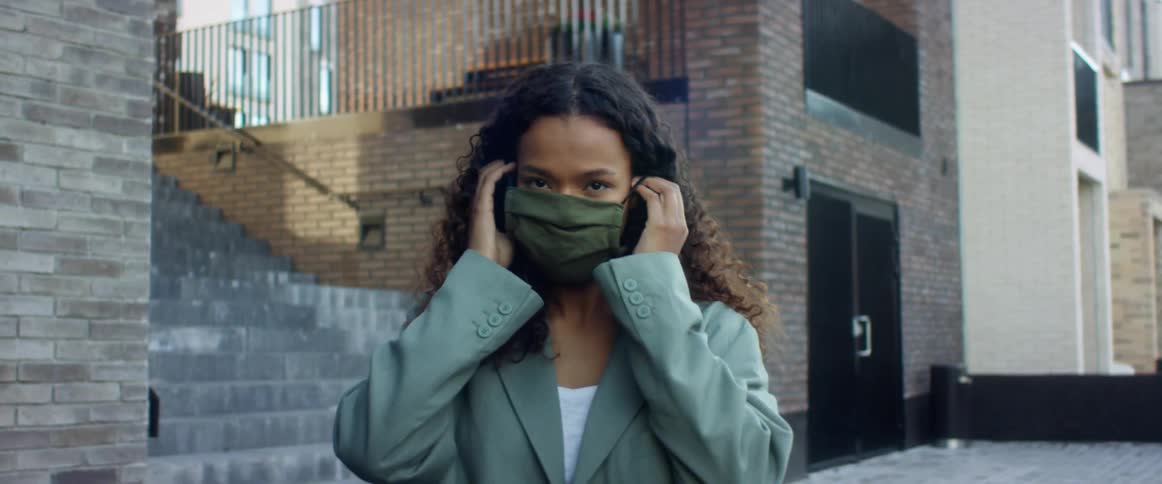 A woman stands outside of a city building and puts on a face mask