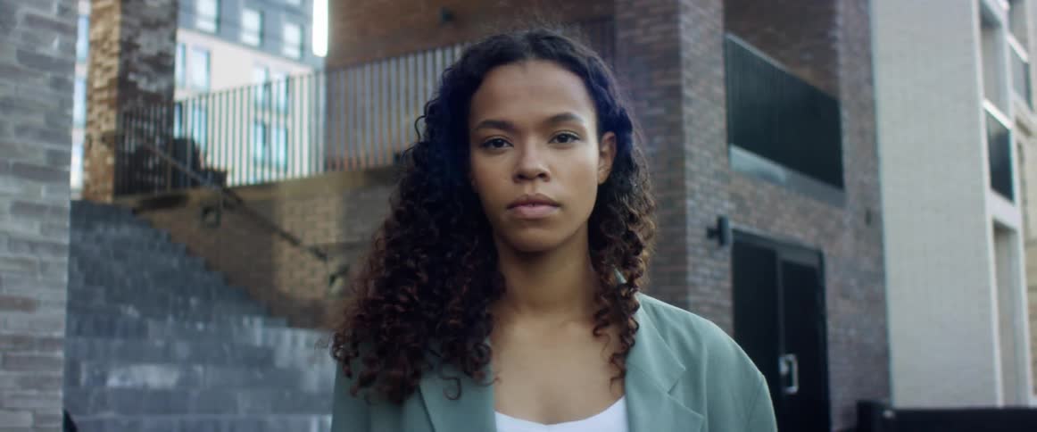 Black woman with curly hair and a blue overcoat faces the camera with eye contact in a street