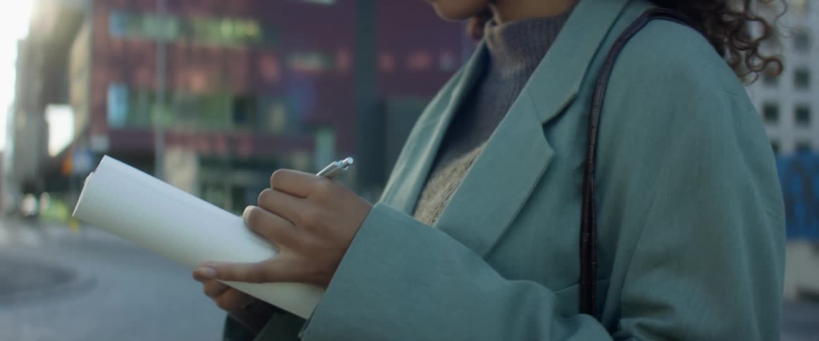 A young woman dressed professionally stands outdoors while writing some notes