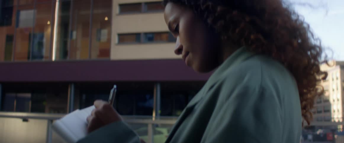 A young black woman writes in a notebook while standing on a bridge outdoors in a metropolis during the day