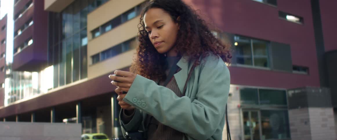 A woman stands outside a large building and pulls her cell phone out of her pocket then smiles as shes looking at it