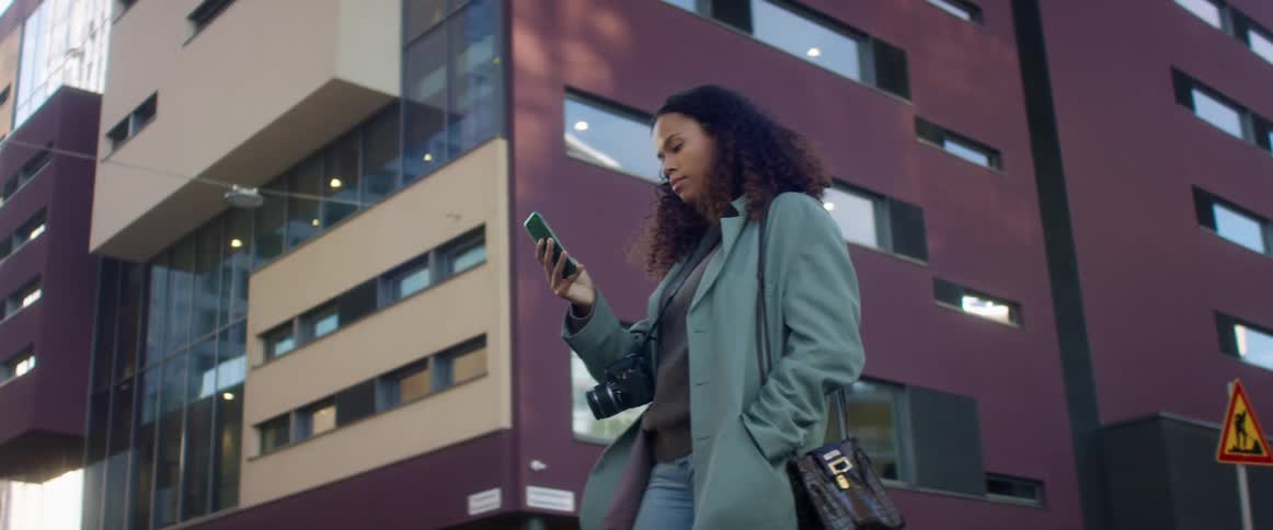 Black woman with curly hair and a blue topcoat uses a smartphone in front of a building in the street