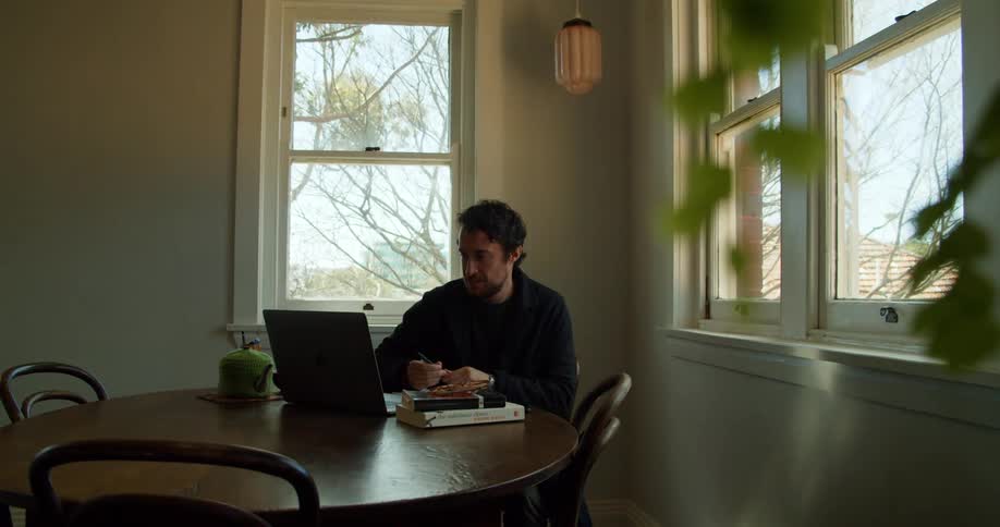 A man sitting at a table in a house uses a laptop to greet someone over video chat