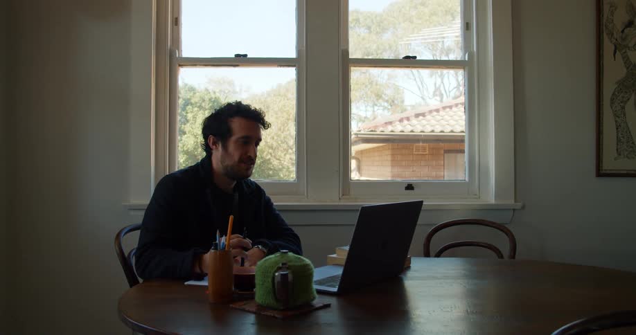 A man sitting at home talks with someone on his laptop and writes something down on paper