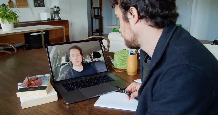 A man sits at a table with a laptop having a video call with another man