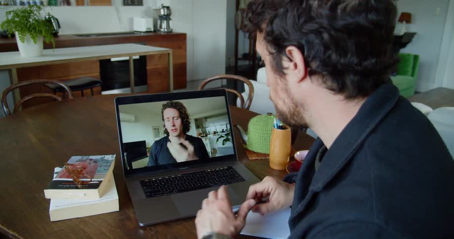 Man with a beard and a black jacket sits at a table and makes a video call on a grey laptop while drinking a cup of tea