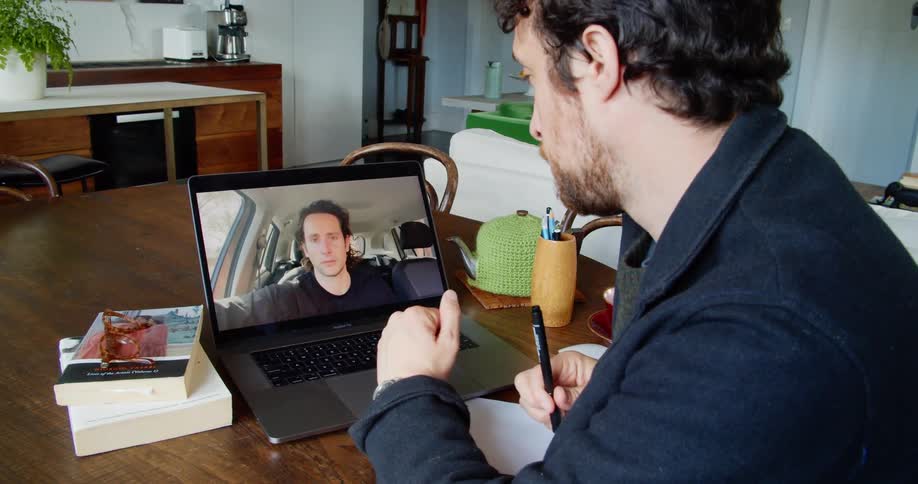 A white man sitting at a table speaking to someone on a video call