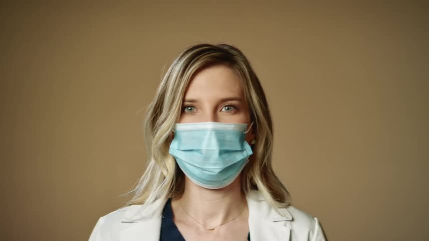 Blonde woman with a blue mask and a white lab coat faces the camera with eye contact