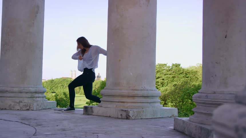 Young fit woman wearing a black body suit dances between the columns of a monument