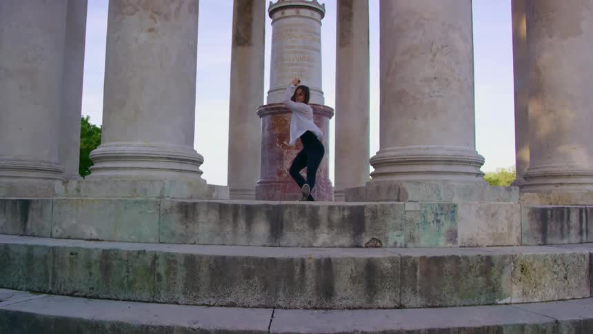 A teenage girl stretches and dances on a platform with large columns