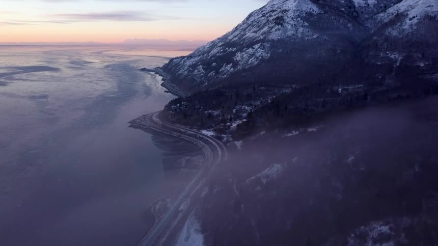 Vehicles drive along a curvy highway sandwiched between the ocean and mountains