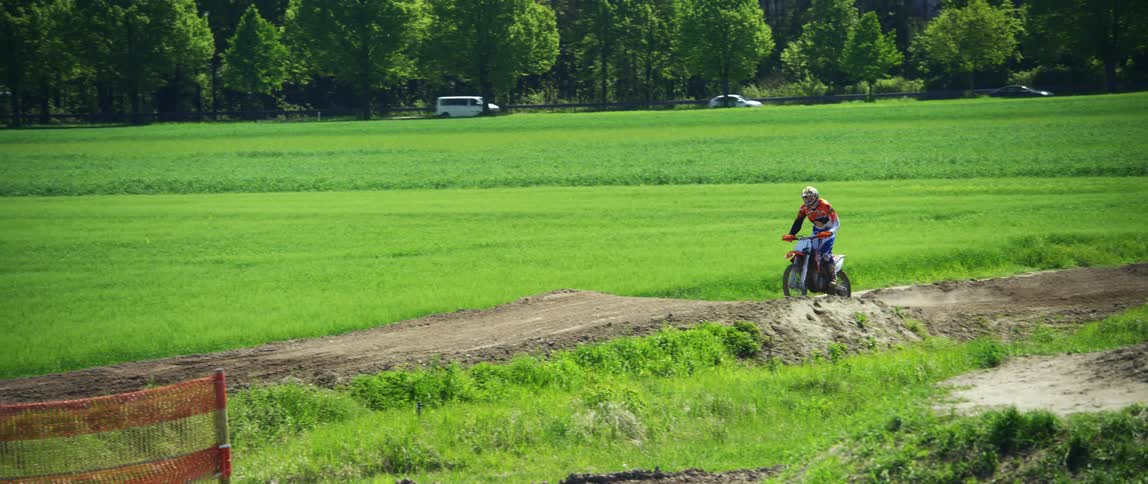A person rides a dirt bike on a rural dirt path with a roadway and vehicles in the distance