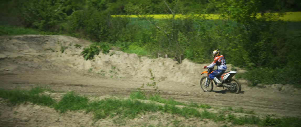 Person rides motorcycle along wet muddy road past building on edge of slope