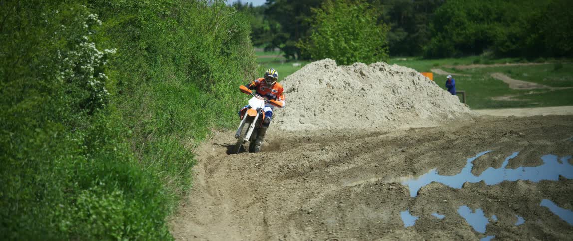 A motocross rider rides along a dirt track riddled with puddles of water on a sunny day