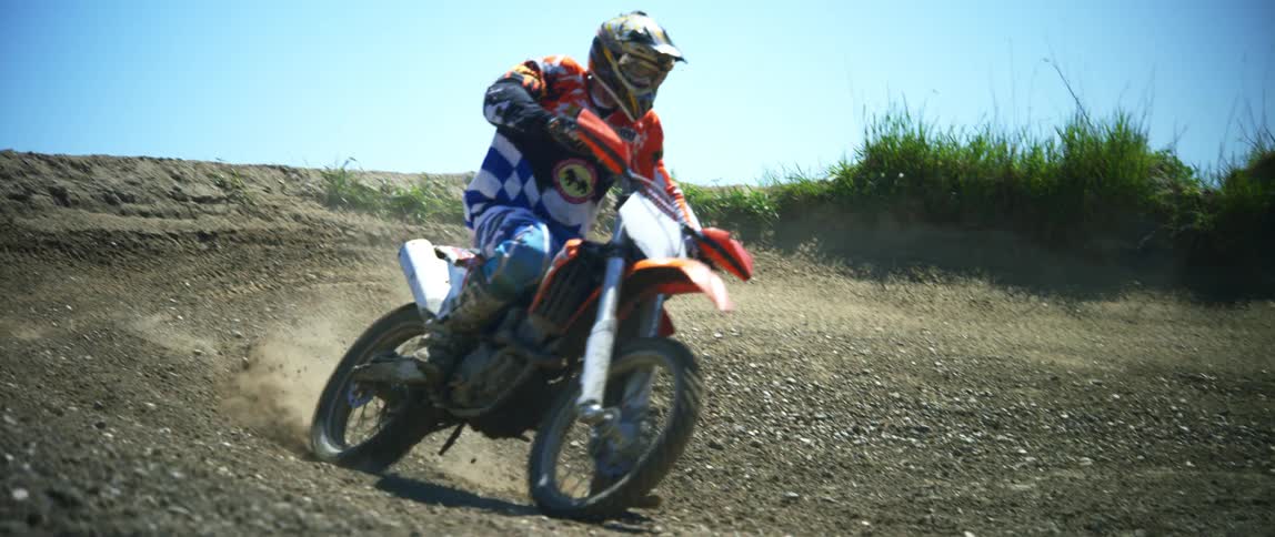 A motorcyclist rides a dirt bike around a sharp bend and off a dirt ramp on an offroad motorcycle course