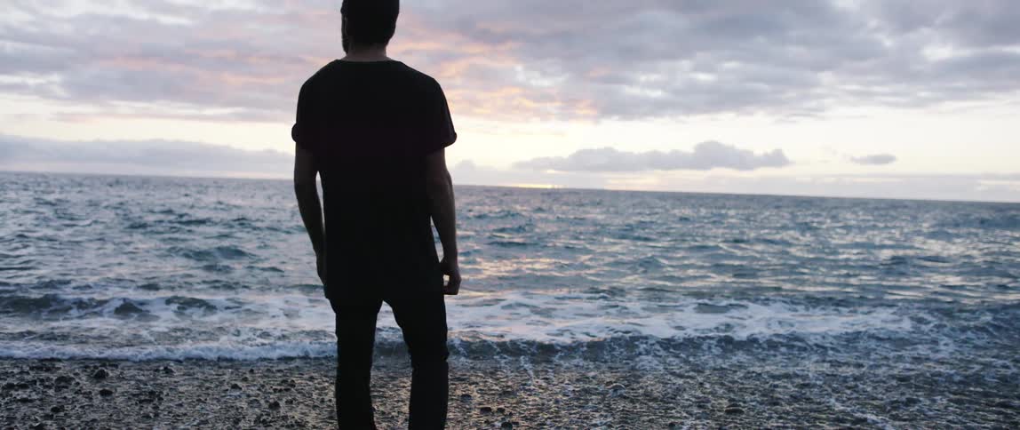 Man with a black t shirt and pants stands on beach with pebbles next to the sea at dusk