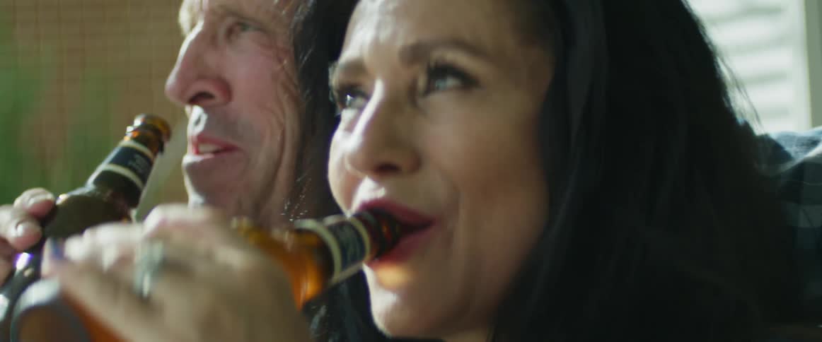 A husband and a wife laugh together while drinking bottles of beer