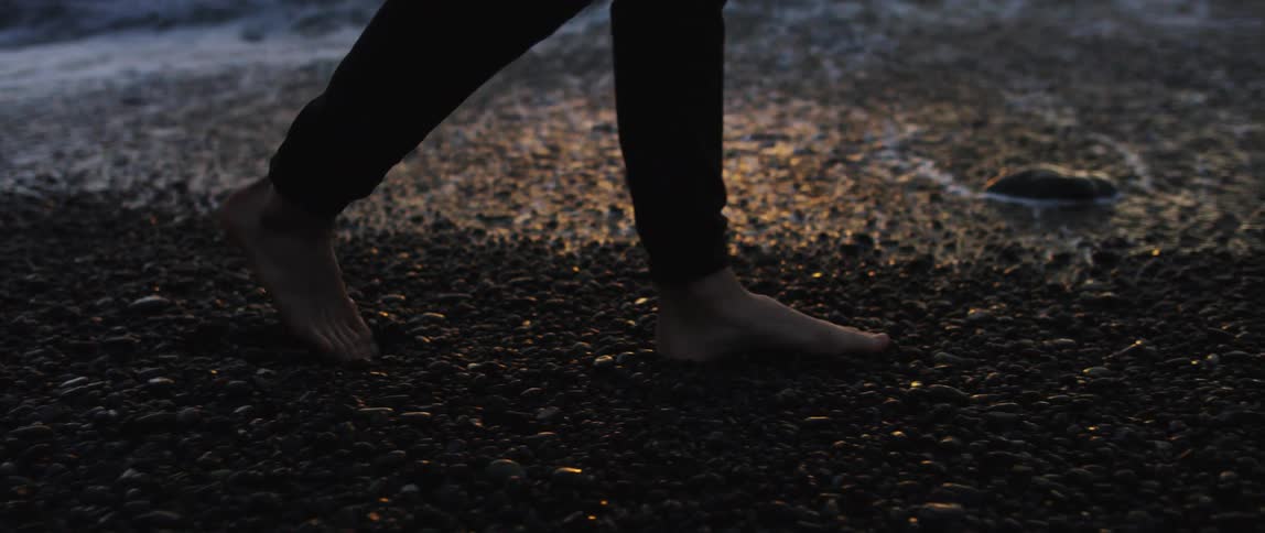 A person walking barefoot on a pebbly beach at sunset
