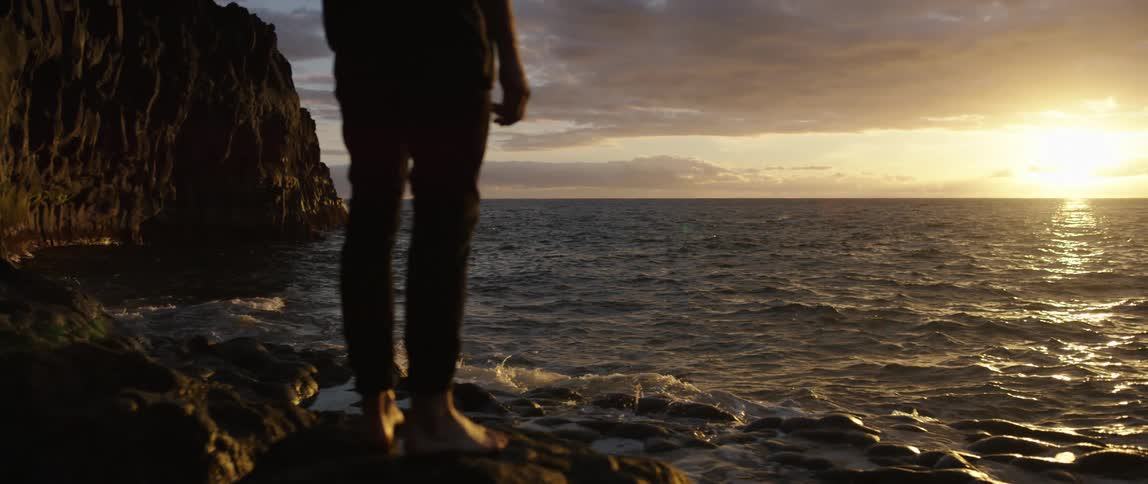 A man standing on rocks on the shore looking out at the sun setting over the water