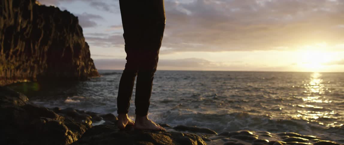 A man stand alone and barefooted on the rocks of a beach at sunset