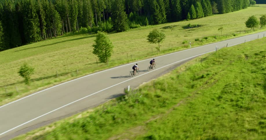 Two cyclists race down a curvy two lane road that runs through a meadow near hills and a forest