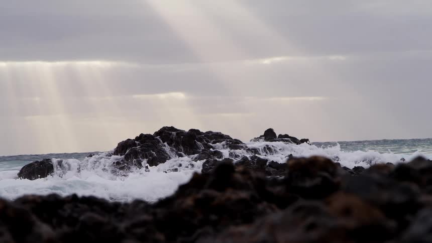 Foamy ocean waves crash against rocks on the shore as the sun shines through the clouds