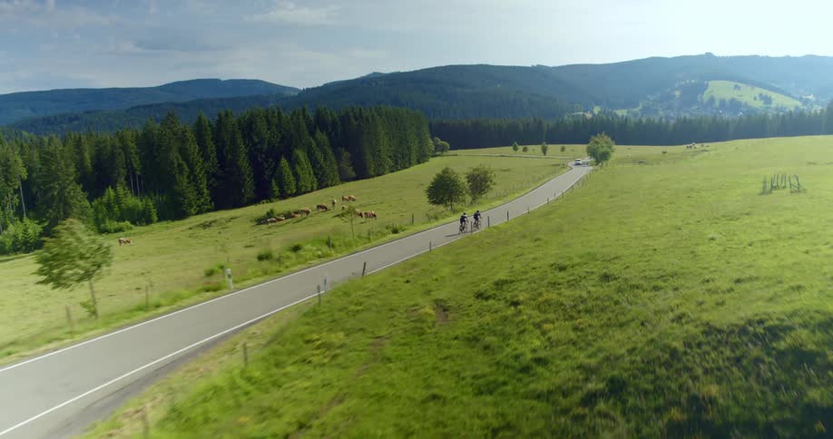 A silver car passes two cyclists racing on a narrow and curvy road in hill country