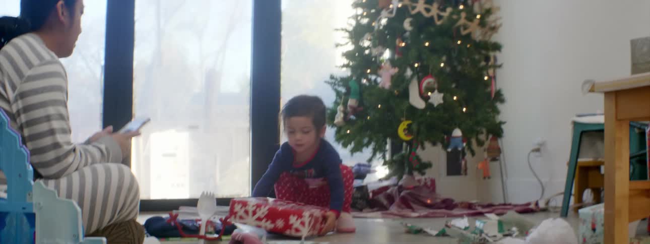 A toddler girl takes a christmas present from under the tree over to the floor near her mother