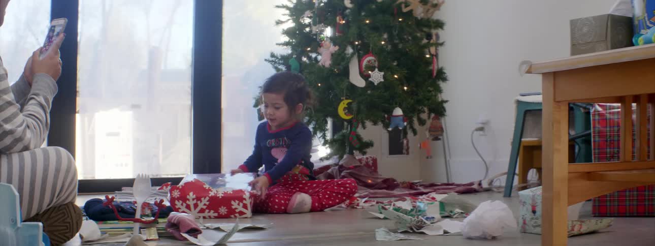 Little girl unwraps christmas presents in the morning while her mother films with her smartphone