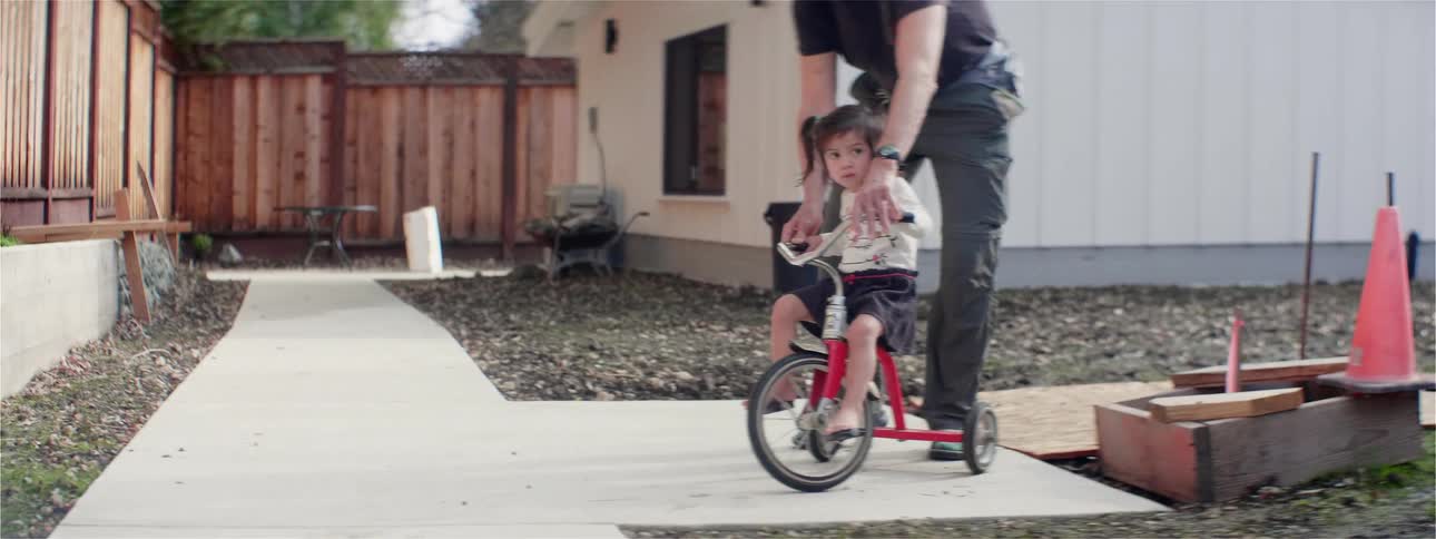 A man pushes a toddler learning to ride a tricycle on a home sidewalk