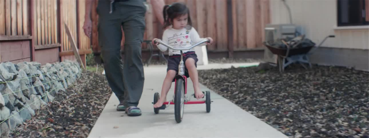 A young toddler girl rides a tricycle on a home sidewalk beside an adult