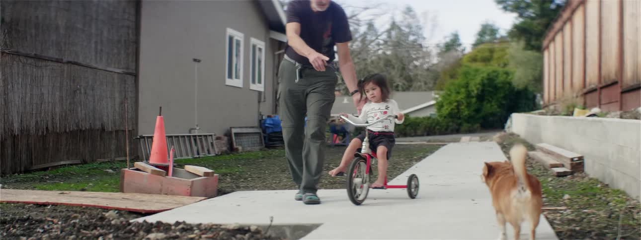 A grandfather helps his toddler granddaughter ride a tricycle as their dog walks toward them