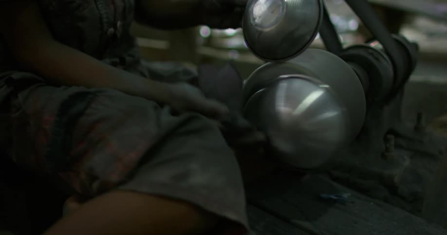 A factory worker uses a towel to hold a metal bowl against a grinding machine