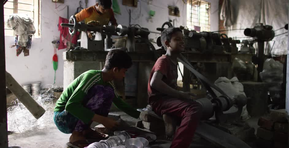 A young boy laughs as he uses a conveyor belt as a massager in a crowded makeshift factory in a village