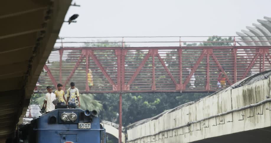 A group of boys walk atop the roof of a train stopped under a bridge