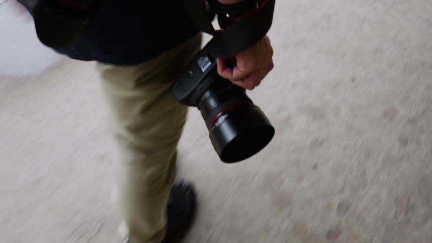 A photographer holds his camera by his right side as he walks down a sidewalk