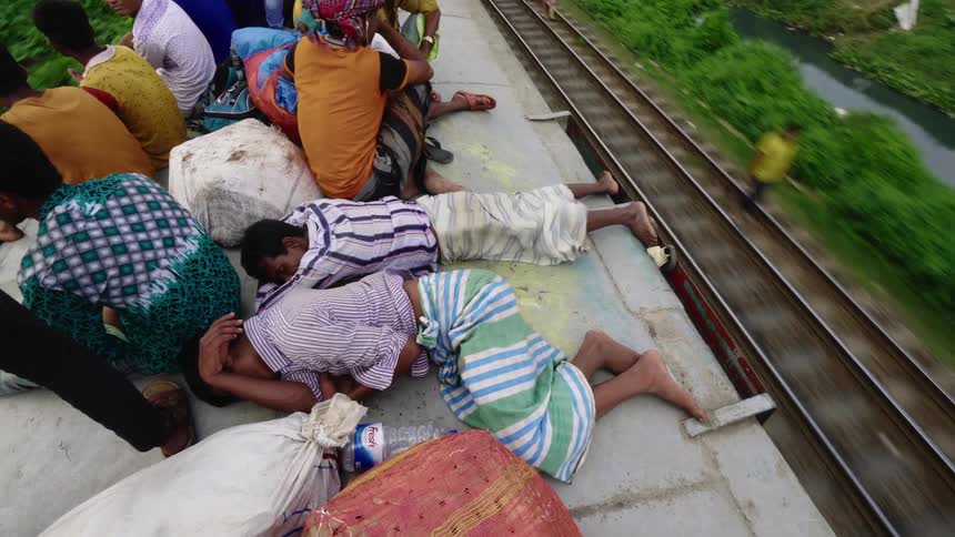 A pair of men sleep on the rooftop of a moving train as other men sit around them