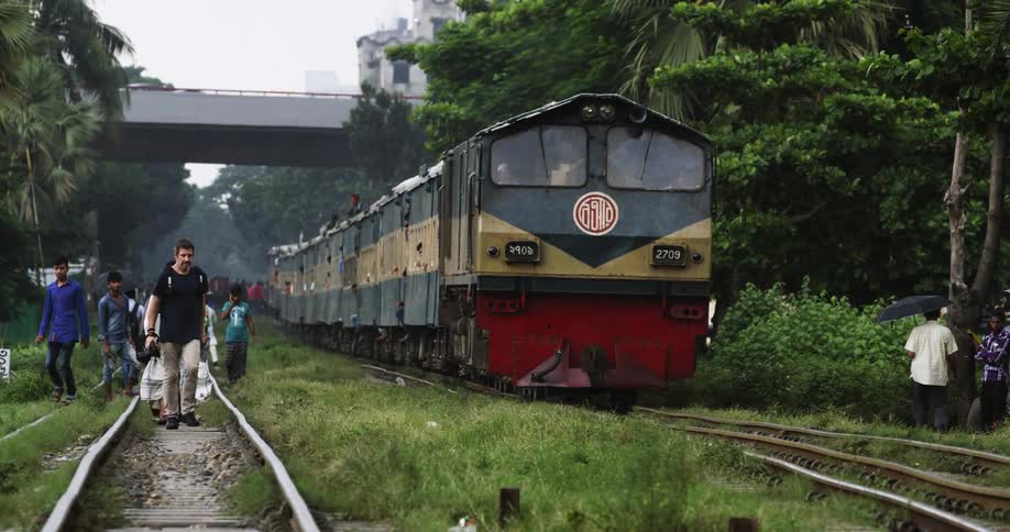 A train moves past a large group of people walking on a track beside it in a rural area