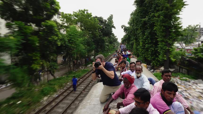 A man with a camera riding on top of a train with other men with the train passing buildings and trees