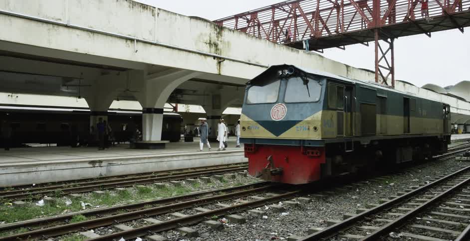 A train pulls up to a station as people walk on the platform and a bridge above