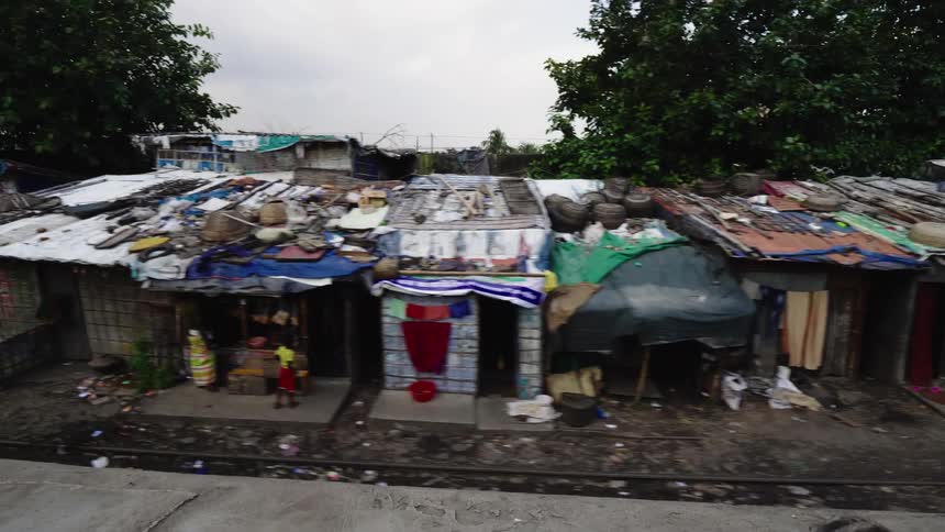 Person on vehicle passes shacks with damaged roofs backed by trees