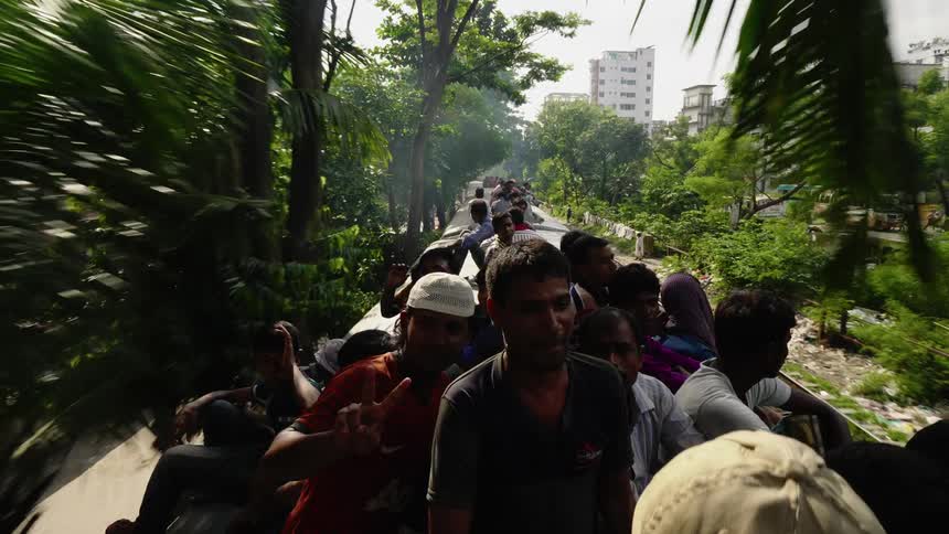 A train with a large group of people on top rides through a primitive village