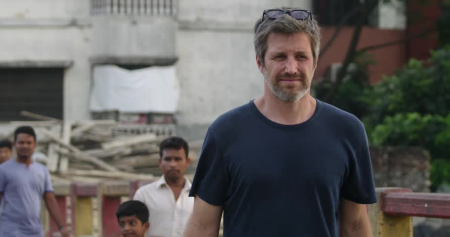 A white man walks down a street with native people in a primitive village