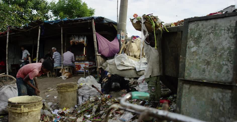 A boy sweeps piles of trash in a field as an older man throws away trash and more people sit in a nearby primitive shop
