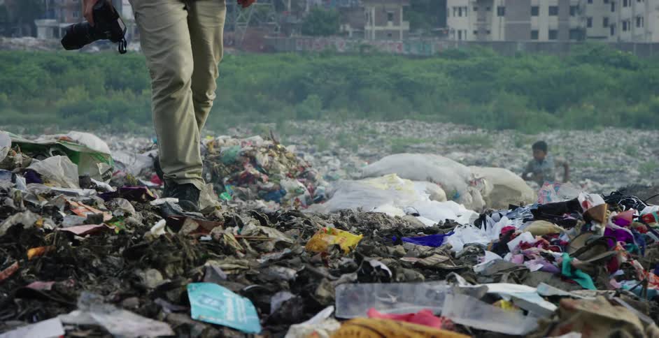 A photographer carefully steps over piles of trash in a landfill while a young boy runs around nearby
