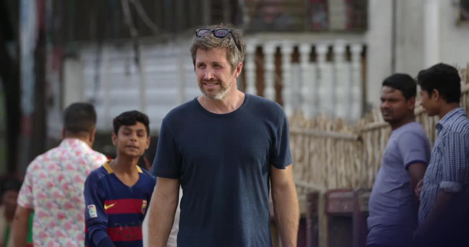 A caucasian man carrying a camera walks down a street in a town in a developing nation