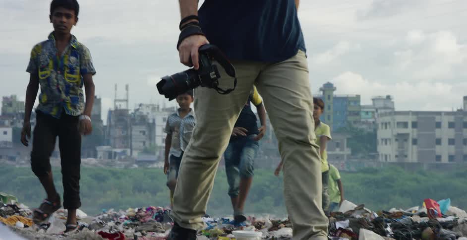 A middled aged photographer walks across a trash heap outside of the slums of a city while followed by a group of children