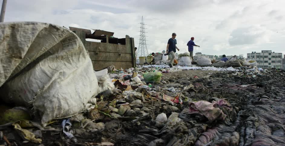 A man with a camera walking with garbage in the foreground and people in the background