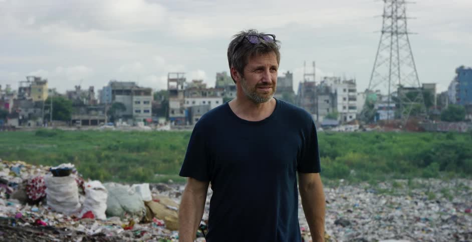 A middle aged man walks over a trash heap in a field outside of a city