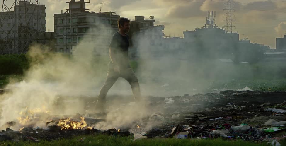 A middled aged man carrying a camera walks through a heap of burning garbage on the outskirts of a rundown city